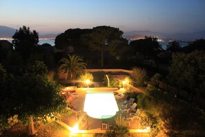 A swimming pool with lounge chairs and a hot tub, surrounded by trees and a view of the city at dusk.