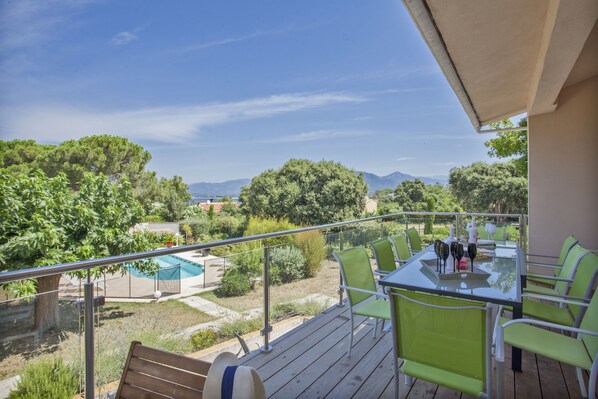 A balcony with a glass table and chairs, overlooking a pool and greenery.