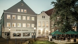 A street view with a building labeled 'Kleiner Amsterdam' featuring a shop on the ground floor, a cafe with outdoor seating, and a tree in the foreground. - Hotel Klein Amsterdam (Friedrichstadt)