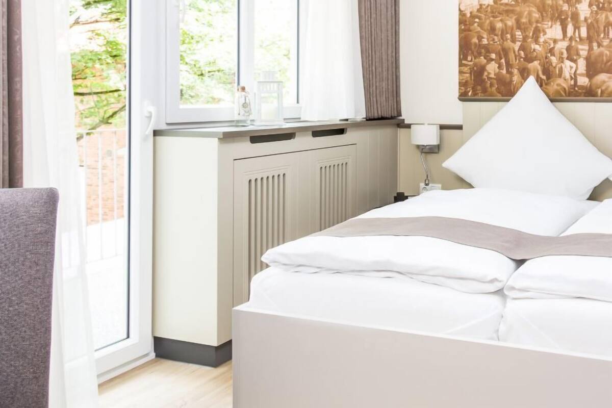 A hotel room with a bed, a window with curtains, a radiator, and a wall-mounted lamp.