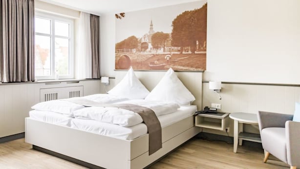 A neatly made bed with white linens and two pillows, a small bedside table with a lamp, and a chair in a modern hotel room with a large window and a cityscape painting on the wall. - Hotel Klein Amsterdam (Friedrichstadt)