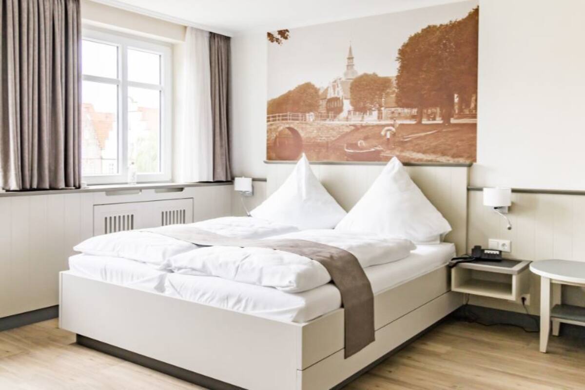 A neatly made bed with white linens and two pillows, a small bedside table with a lamp, and a chair in a modern hotel room with a large window and a cityscape painting on the wall.