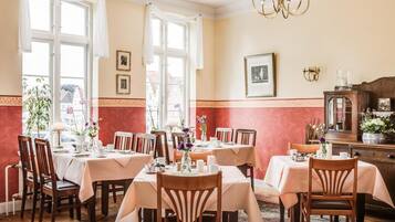 Un comedor con varias mesas dispuestas para comensales, amplias ventanas con cortinas y un candelabro.