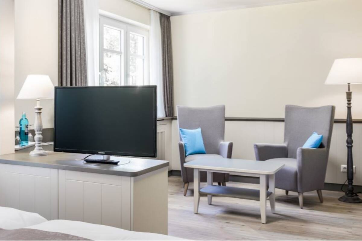 A hotel room with a bed, a TV on a stand, a small table, two armchairs, and a floor lamp.