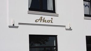 A building facade with the name "Ahoi" in gold lettering, a security camera, and a window with a curtain. - Boutique Pension Ahoi (Wesselburenerkoog)