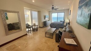 A spacious hotel room with a balcony, a large bed, a desk with a chair, and a seating area with a sofa and armchairs.
