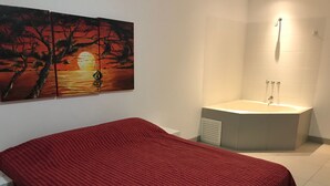 Une chambre à coucher avec une couvre-lit rouge, un grand tableau représentant un coucher de soleil et une baignoire équipée d’un pommeau de douche.