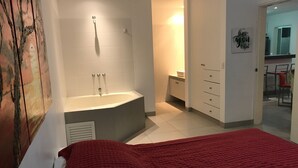 Une chambre avec un grand lit, une salle de bain avec une baignoire, et un coin cuisine visible à travers une porte ouverte.