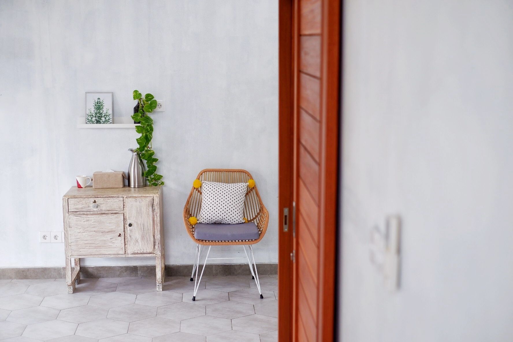 A room with a wooden dresser, a wicker chair with a cushion, a potted plant, and a framed picture on the wall.