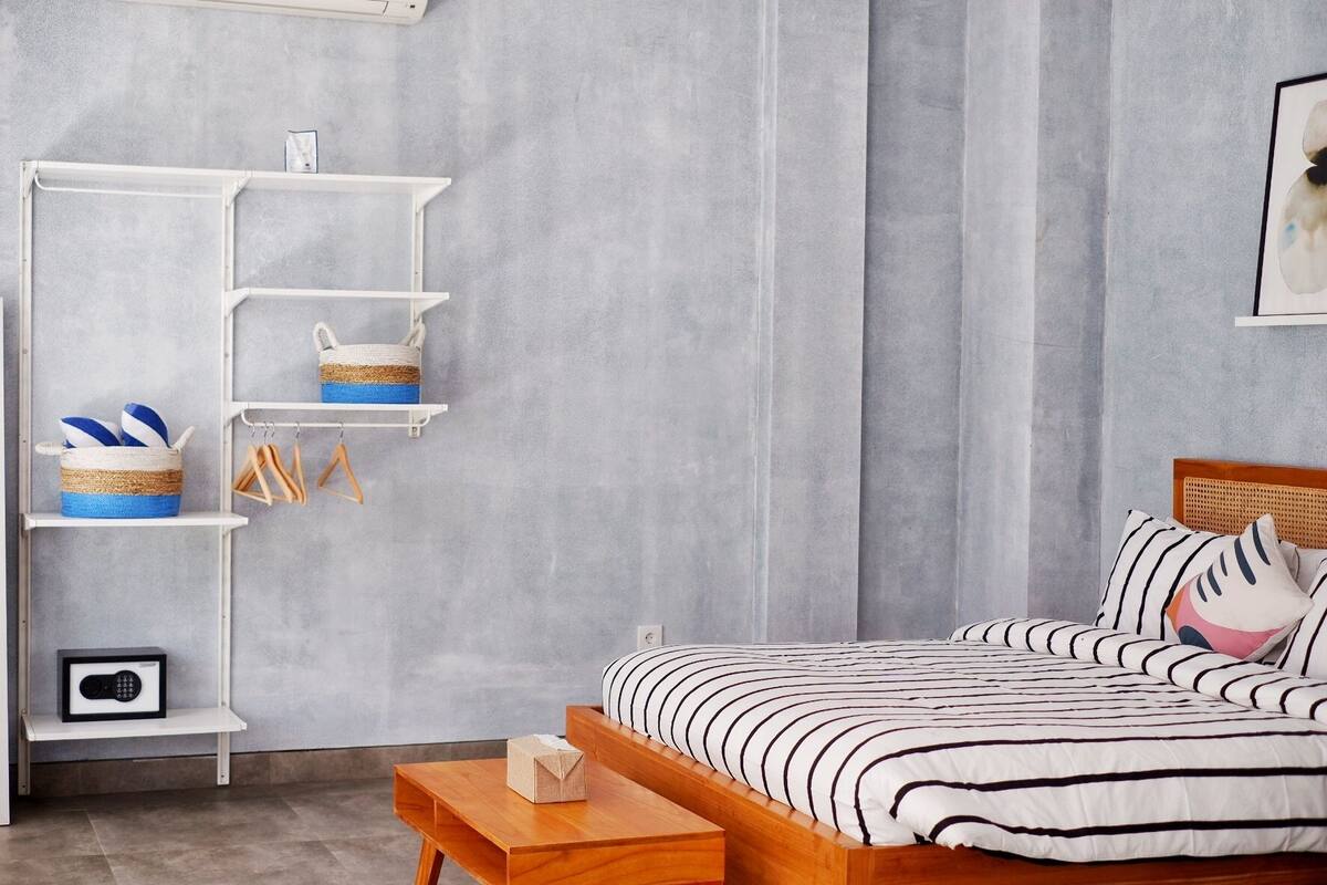 A bedroom with a bed, a wooden nightstand, a white shelving unit with baskets and hangers, and a grey textured wall.