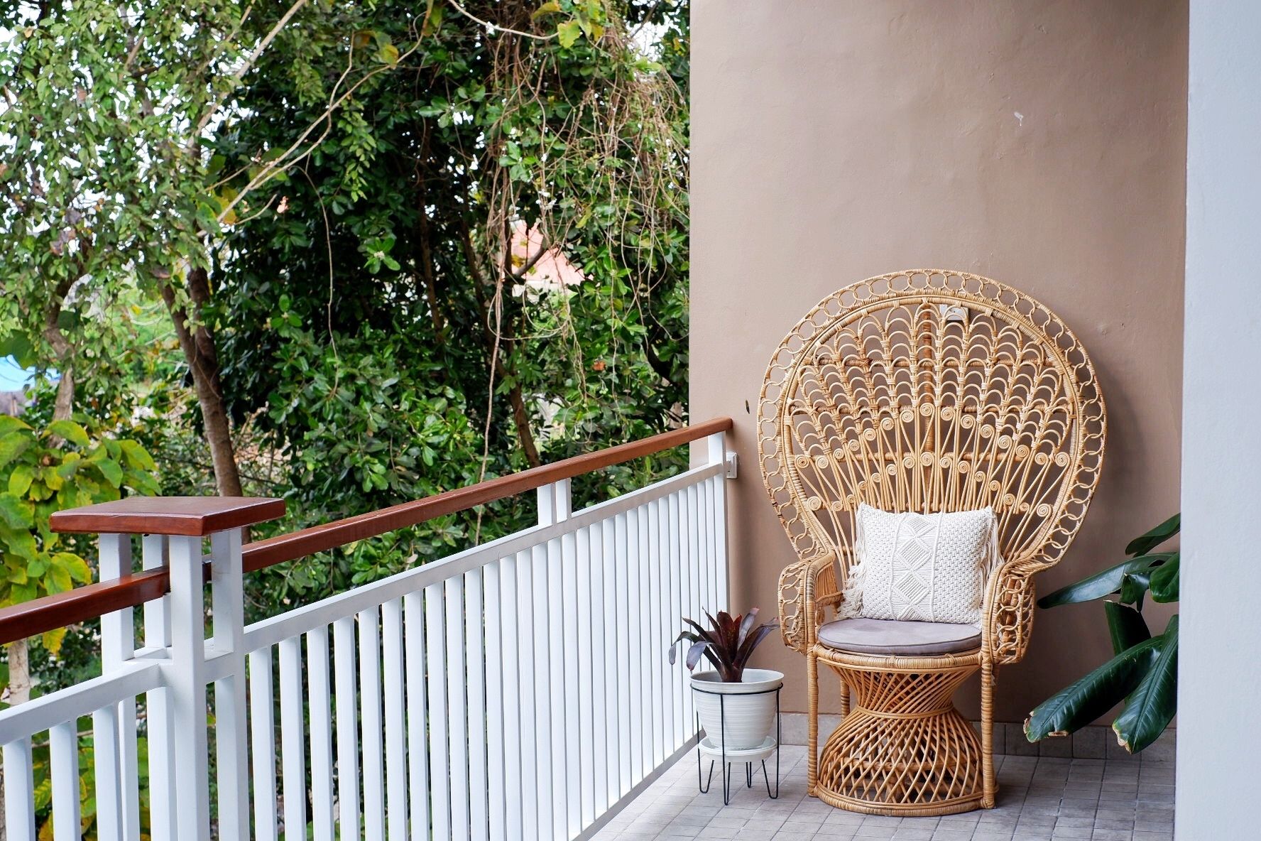 A wicker chair with a cushion on a balcony with a white railing, surrounded by greenery.