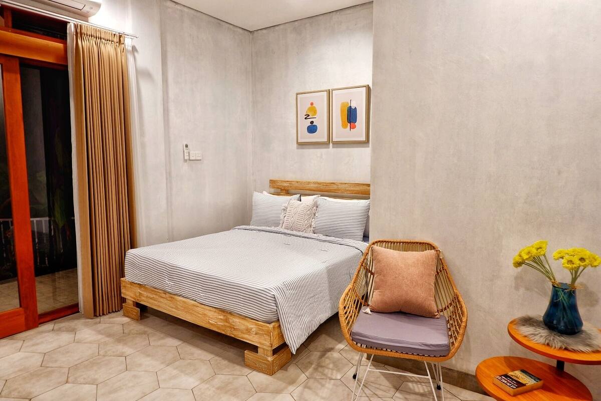 A bedroom with a wooden bed, a wicker chair, a small table with a vase of flowers, and a sliding glass door.