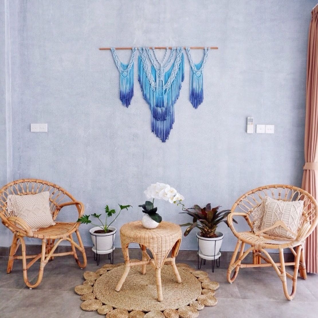 Une pièce comprenant deux fauteuils en osier, une petite table ronde ornée d’une orchidée blanche et une décoration murale en macramé.