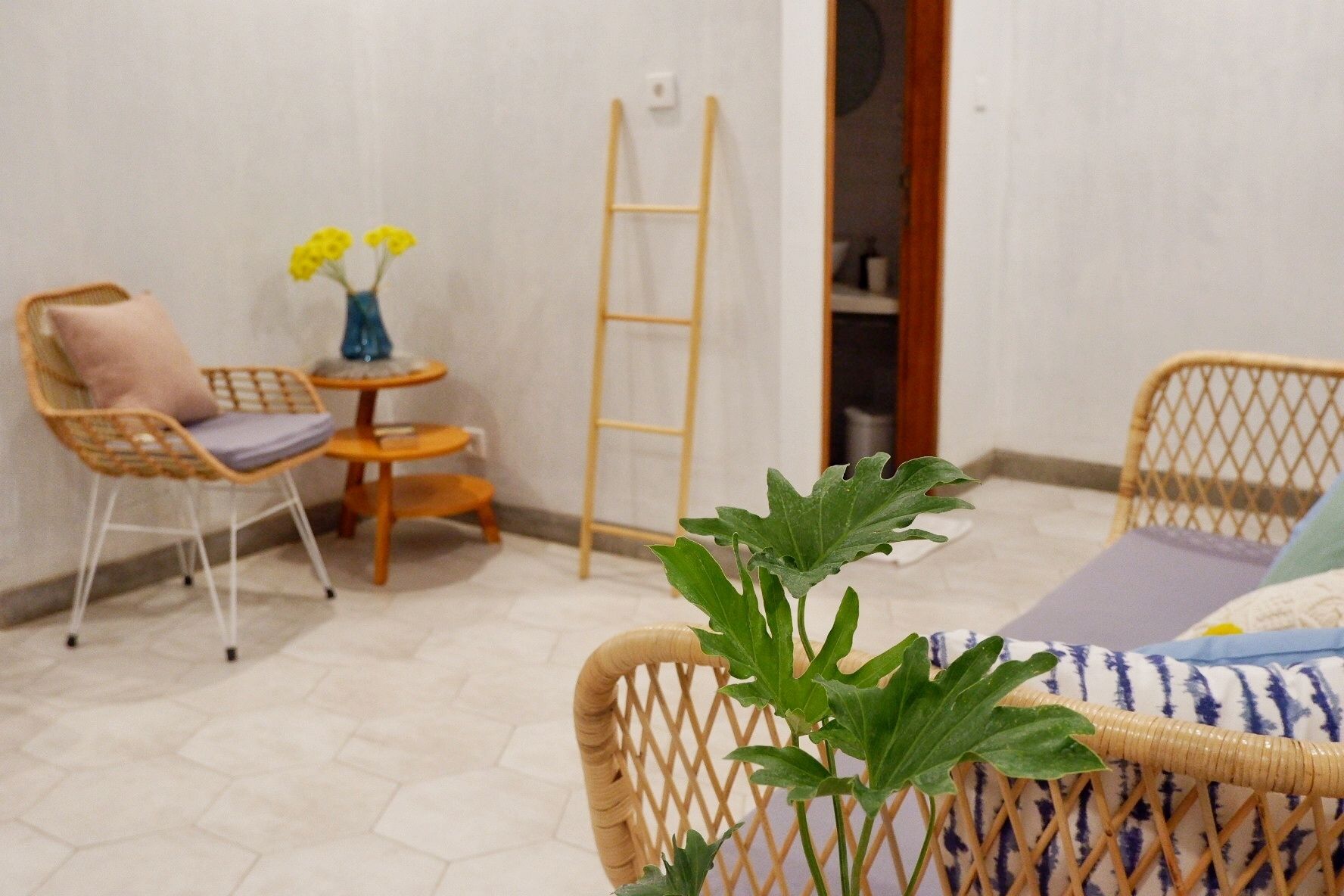 A small, cozy room with a wicker chair, a small table with a vase of yellow flowers, and a ladder leaning against the wall.