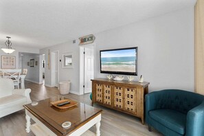 Un salon avec un téléviseur diffusant une scène de plage, une table basse en osier, un canapé bleu et un meuble en bois orné d’objets décoratifs.