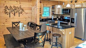 A wooden cabin interior with a kitchen area featuring a stainless steel refrigerator, a microwave, and a coffee maker. There is a dining table with chairs and a small bar stool.