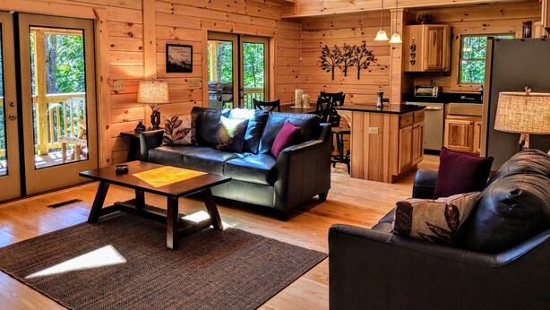 A cozy cabin interior with a wooden floor, a sofa, a coffee table, and a kitchen area with a refrigerator, stove, and cabinets.