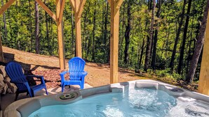A hot tub with two blue Adirondack chairs, surrounded by a wooden pergola and a forested area.