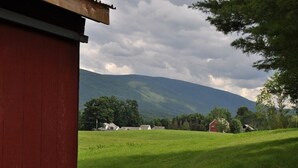Un granero rojo en un entorno rural con un campo cubierto de hierba, algunas casas y una montaña al fondo.
