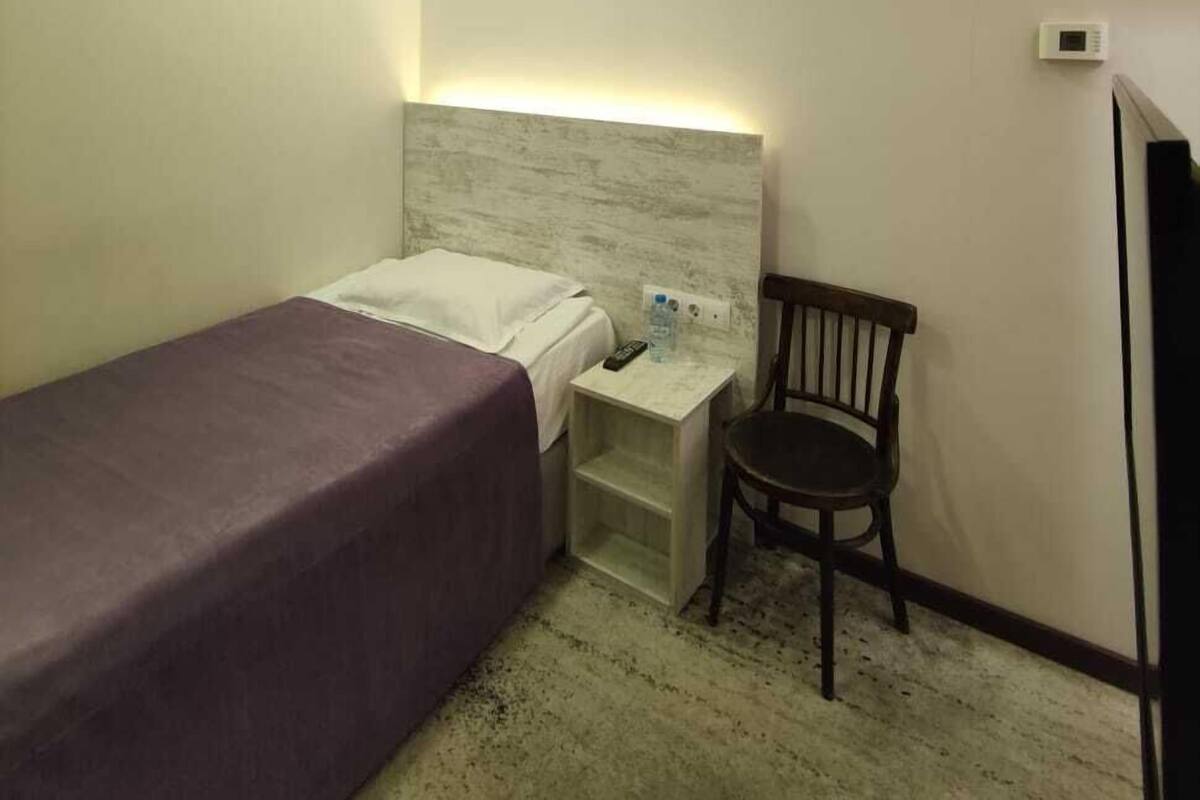 A small, single-bed room with a headboard, a nightstand, a chair, and a wall-mounted thermostat.