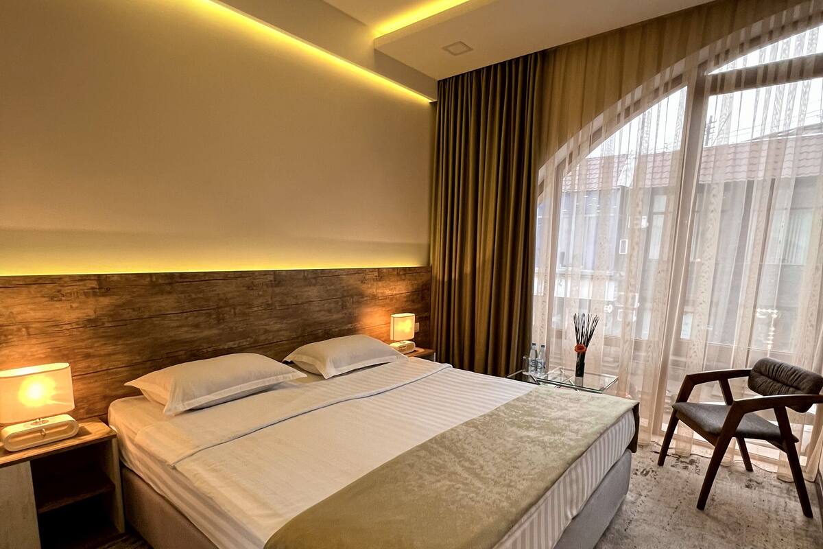 A modern hotel room with a large bed, bedside lamps, a wooden headboard, a glass table with a vase, and a view of the outside through sheer curtains.