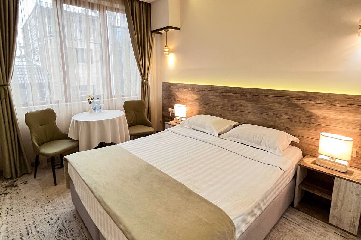 A hotel room with a large bed, a round table, two chairs, a bedside table with a lamp, and a view of buildings through sheer curtains.