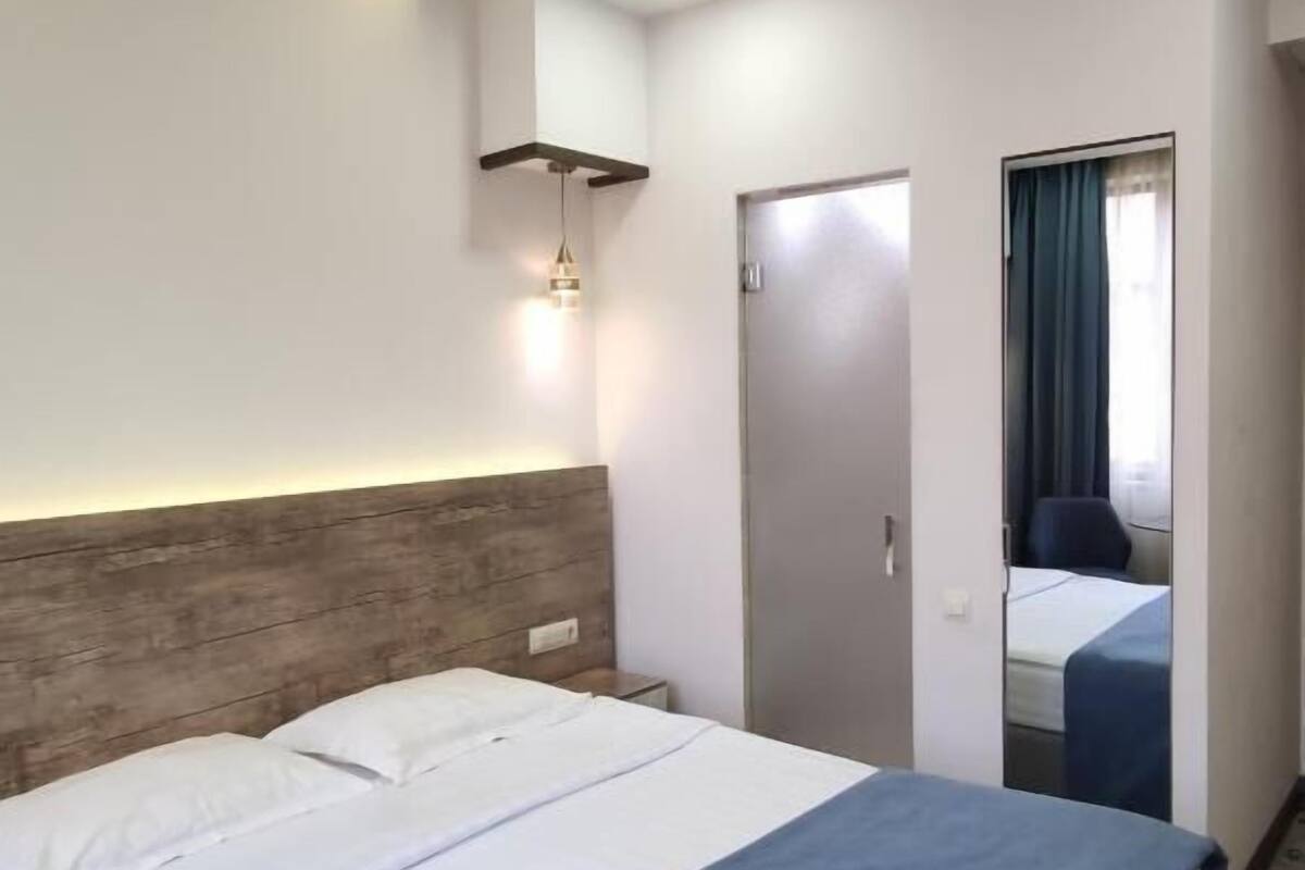 A hotel room with a bed, a wooden headboard, a nightstand, a wall-mounted light, and a door leading to another room.