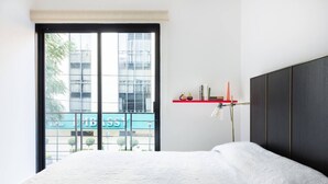 Une chambre avec une grande fenêtre, un lit, une table de chevet avec une lampe, et une étagère rouge contenant des livres et des objets décoratifs.