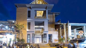 A multi-story building with a modern design, featuring a prominent entrance with steps, a balcony on the upper floor, and a parking area with motorcycles and a car.