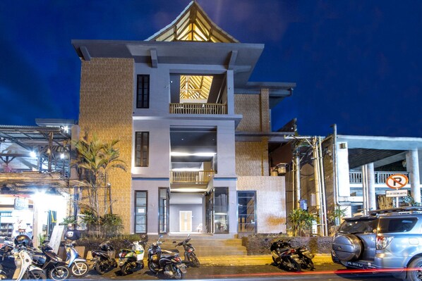 Un edificio de varios pisos con diseño moderno, que destaca por una entrada con escalones, un balcón en el nivel superior y un estacionamiento con motocicletas y un coche.