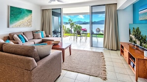 A modern living room with a sofa, coffee table, and a TV stand. It features a large sliding glass door leading to a balcony with a view of the sea and mountains. - Frangipani 8 - ground floor apartment (Hamilton Island)
