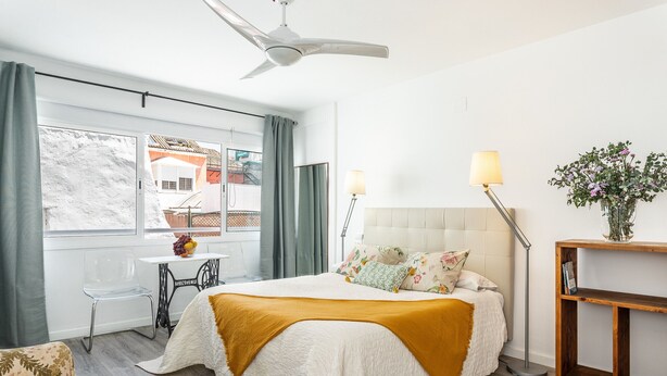 Quarto moderno com cama, escrivaninha e cadeira. Há uma janela com cortinas, um ventilador de teto e uma mesa de cabeceira com abajur.