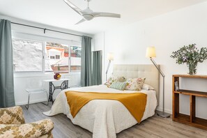A modern bedroom with a bed, a desk, and a chair. There is a window with curtains, a ceiling fan, and a bedside table with a lamp.