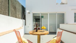 Uma mesa de madeira redonda com duas cadeiras laranja, uma garrafa de vinho e um copo de vinho tinto numa varanda com uma parede branca e uma porta de vidro que dá para um espaço interior.