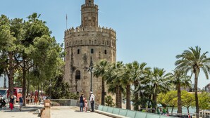 Uma torre histórica com cúpula e merlões, cercada por palmeiras e uma passarela para pedestres.