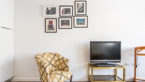Uma sala de estar com uma poltrona estampada, uma TV de tela plana em um suporte dourado e uma parede com quadros.