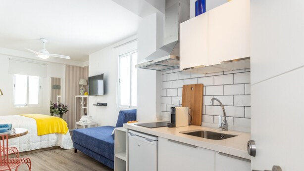 Quarto de hotel compacto com cozinha compacta, cama com cobertor amarelo, escrivaninha com cadeira e ventoinha no teto.