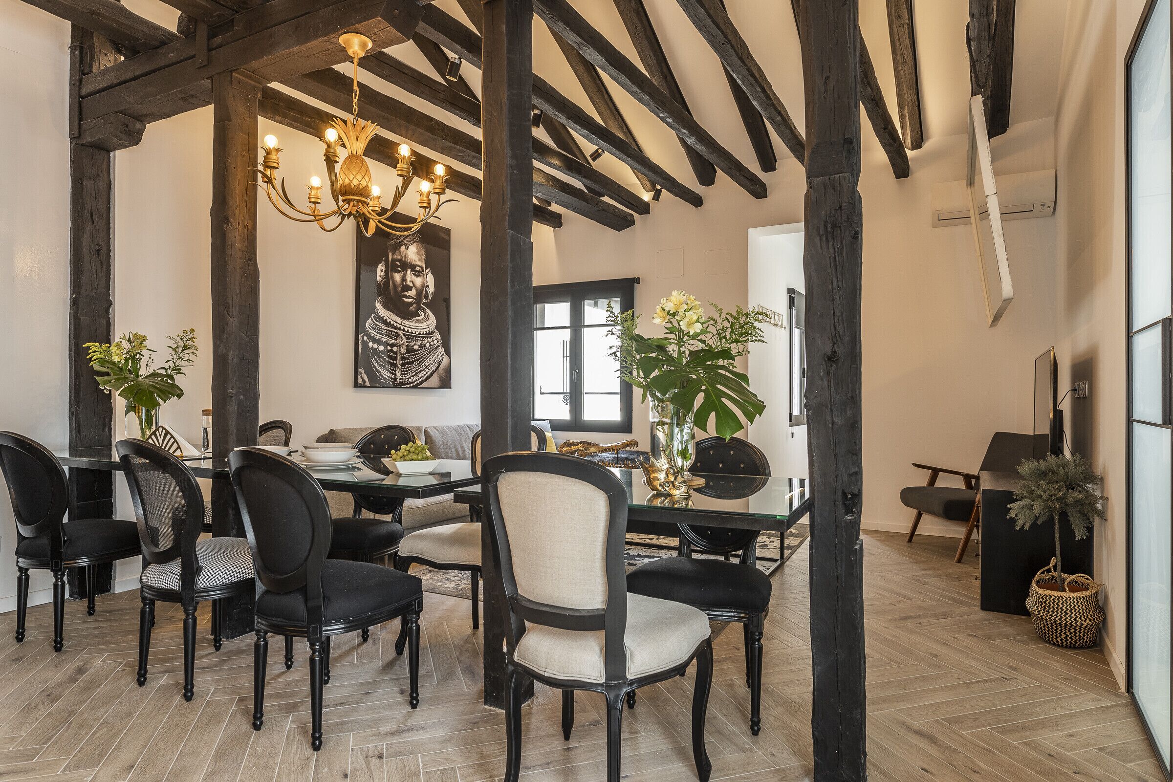 A dining area with a wooden ceiling, black chairs, a chandelier, and a large painting on the wall.