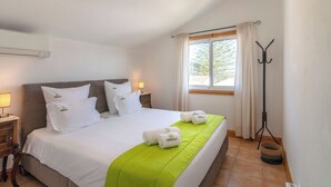 En pænt redt seng med hvide sengetøj og grønne detaljer, et natbord med lampe, et vindue med gardiner og en gulvlampe.