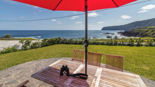 En træterrasse med en rød parasol, to træstole og kikkert, med udsigt over et kystlandskab med klipper og hav.