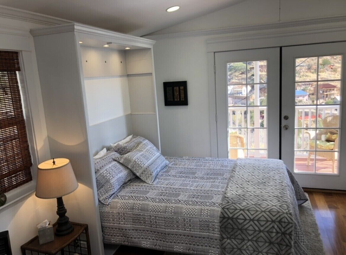 A bedroom with a bed, a bedside table with a lamp, a window with blinds, and a door leading to an outdoor area.