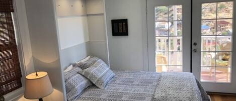 A bedroom with a bed, a bedside table with a lamp, a window with blinds, and a door leading to an outdoor area.