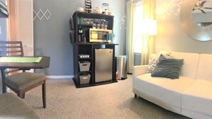 A living room with a white sofa, a dining table with chairs, a black bookshelf, and a small refrigerator. - Relax in Your Own Private Suite in Charleston's James Island! (Charleston)