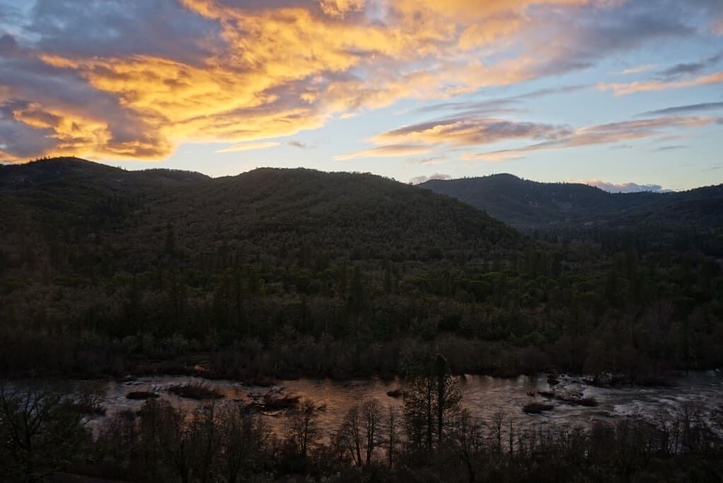 A mountainous landscape with a river in the foreground and a sunset in the background.