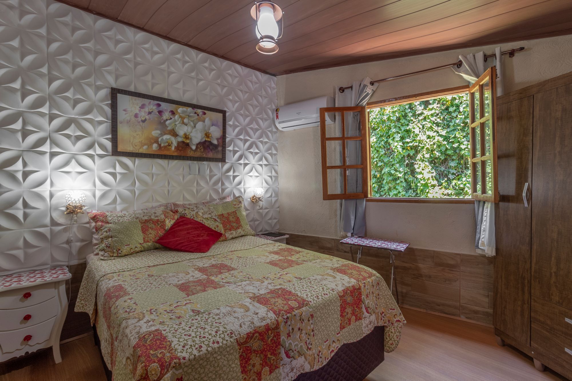 Un dormitorio con cama, mesita de noche, ventana con contraventanas abiertas y pared con diseño geométrico.