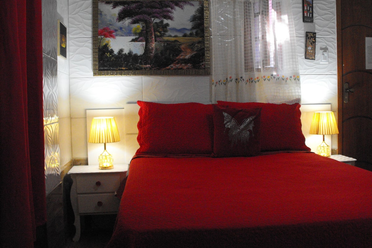 Un dormitorio con una colcha roja, dos lámparas de noche, una mesita de noche y un cuadro en la pared.