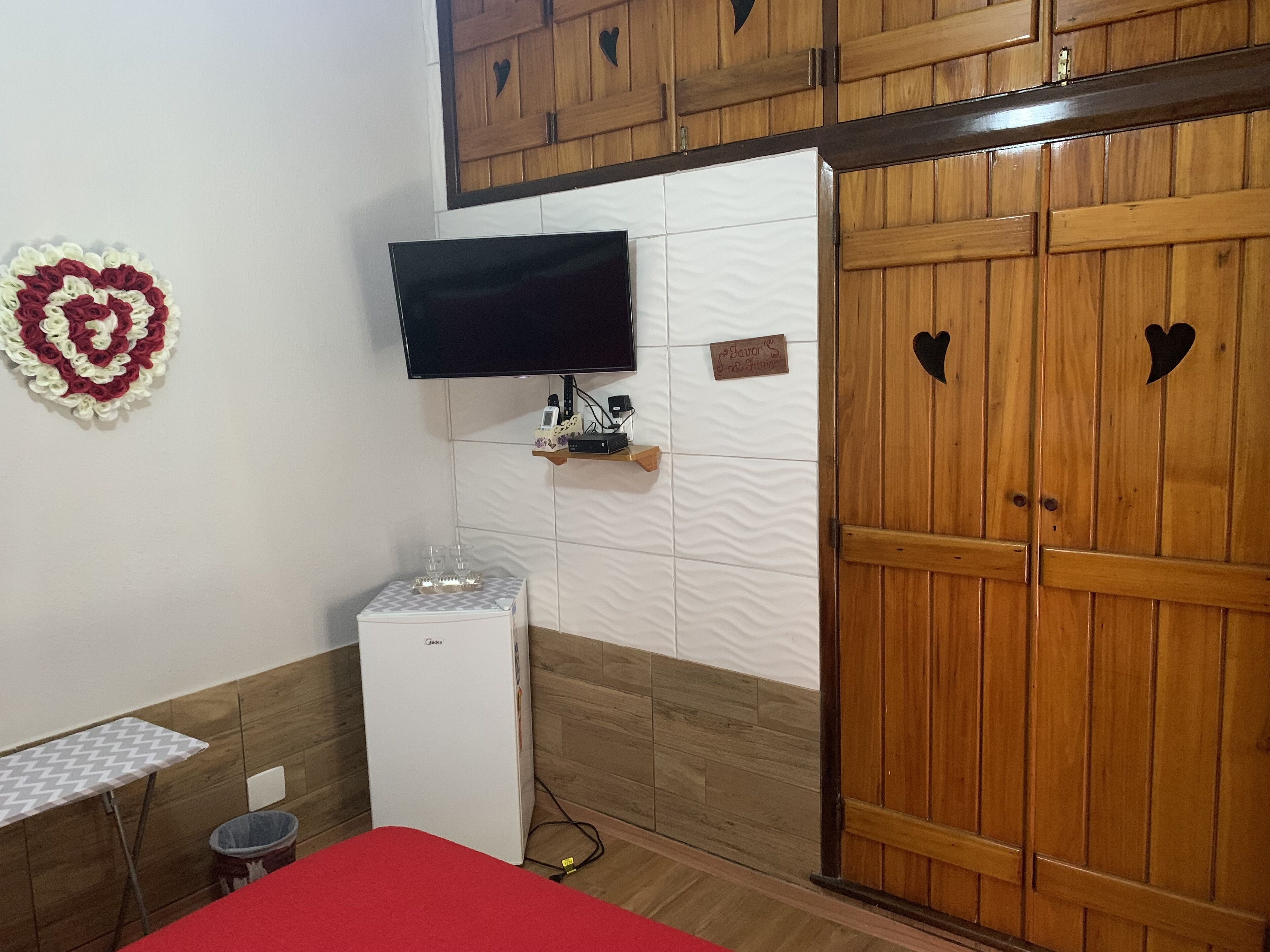 A room with a wooden wardrobe, a television mounted on the wall, a small refrigerator, and a heart-shaped decoration on the wall.