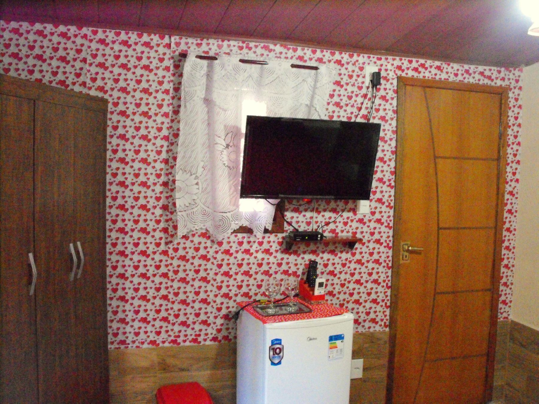 Ein Zimmer mit an der Wand befestigtem Fernseher, einem Kühlschrank, einer Holztür und einem Kleiderschrank.