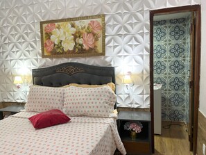 A bedroom with a tufted headboard, a painting of flowers, two bedside lamps, patterned bedding, a red pillow, and a mirror reflecting patterned tiles. - Pousada Cascata dos Amores (Teresópolis)