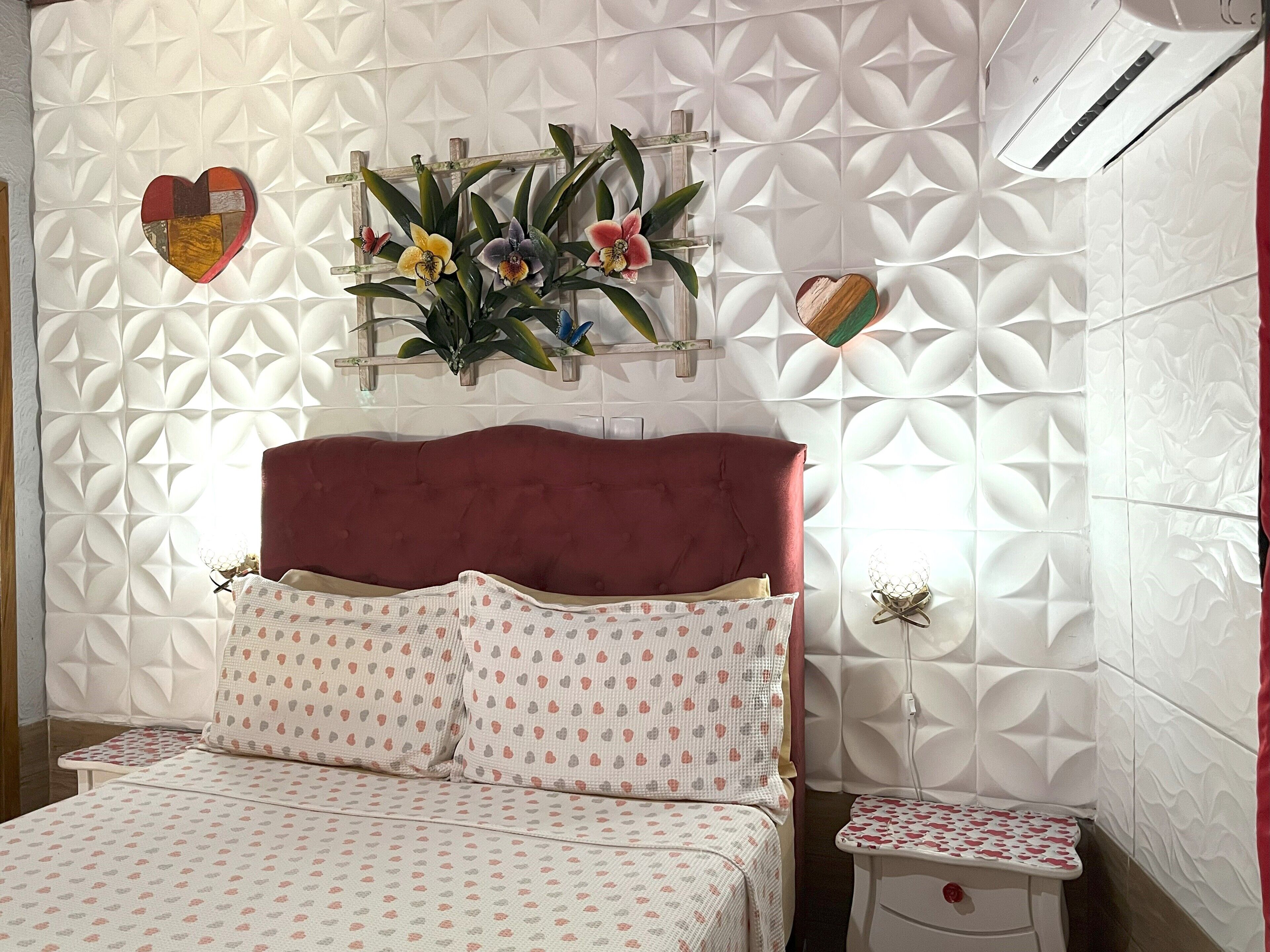 A bedroom with a red upholstered headboard, a bed with patterned bedding, a small bedside table, and a wall-mounted air conditioner.